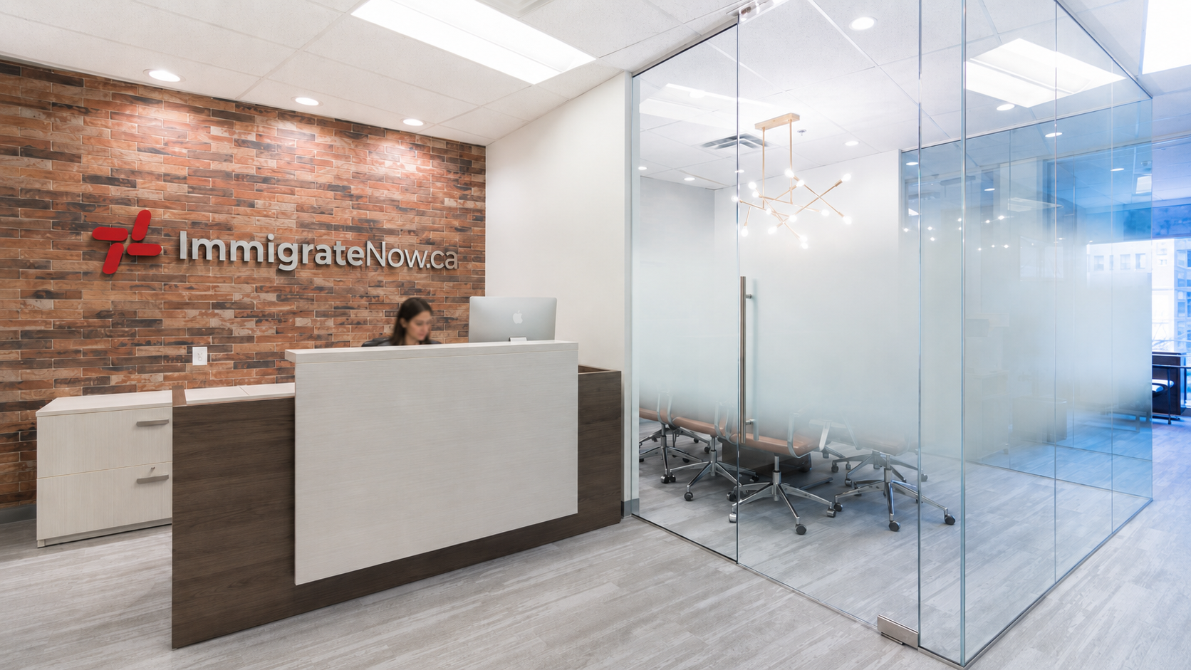 Immigrate Now reception with the Immigrate Now.ca signage on a wood-panelled wall and a glass-walled meeting room in the background